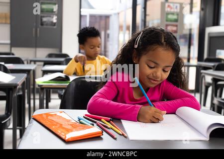 Fille biraciale avec les cheveux bruns bouclés est dans une salle de classe d'école, garçon afro-américain en arrière-plan Banque D'Images