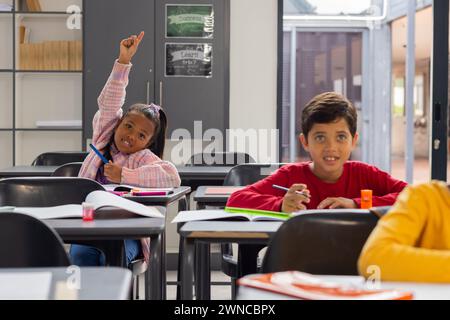 Fille biraciale dans une chemise à carreaux rose lève sa main avec impatience dans une salle de classe d'école Banque D'Images