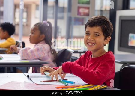 Garçon biracial avec un sourire brillant est assis à un bureau dans une salle de classe d'école, cheveux foncés soigneusement peignés Banque D'Images