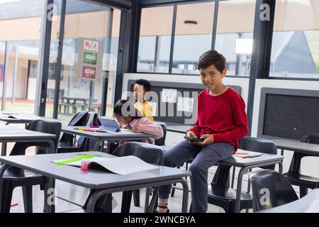 Garçon biracial dans un pull rouge tient un téléphone intelligent dans une salle de classe avec espace de copie Banque D'Images