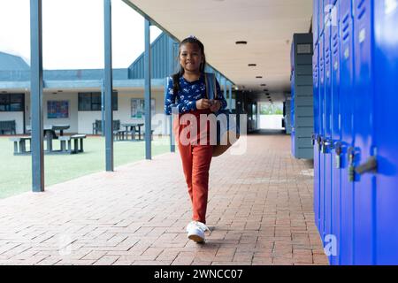 Fille biraciale avec un sac à dos se tient près de casiers bleus à l'école Banque D'Images