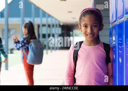 Fille biraciale sourit par des casiers, une autre fille en arrière-plan à l'école. Banque D'Images