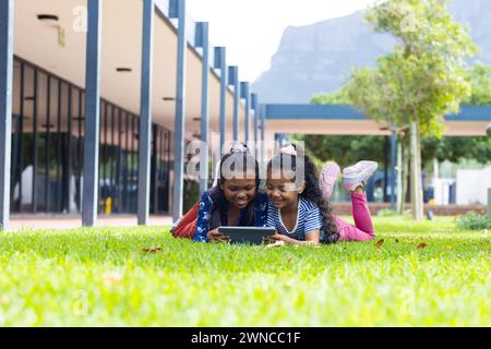 Deux filles biraciales sont engrossées dans une tablette à l'extérieur à l'école, allongées sur l'herbe Banque D'Images