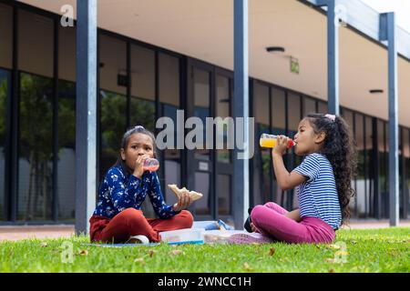 Deux filles biraciales profitent du déjeuner à l'école, assises sur l'herbe avec des sandwichs et du jus Banque D'Images