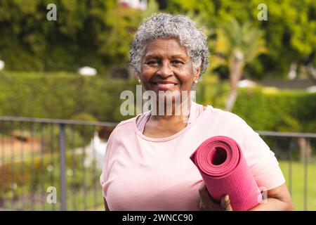 Femme biraciale senior avec les cheveux gris tient un tapis de yoga rose à l'extérieur Banque D'Images