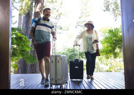 Couple senior biracial en tenue de vacances décontractée marchant avec des valises Banque D'Images