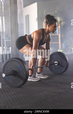 Jeune femme biraciale forte forme soulevant des poids dans un gymnase, axée sur son exercice de soulevé de terre Banque D'Images