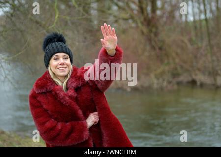 Belle dame blonde portant une veste rouge et des vagues de chapeau de boule noir à quelqu'un dans la distance Banque D'Images