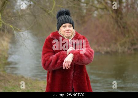 Jolie dame blonde portant une veste rouge et un chapeau noir bouffant faisant un visage drôle et pointant vers sa montre disant que quelqu'un est en retard Banque D'Images