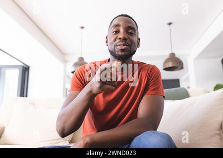 Homme afro-américain dans une chemise rouge tient une télécommande et se détendant sur un canapé en appel vidéo Banque D'Images