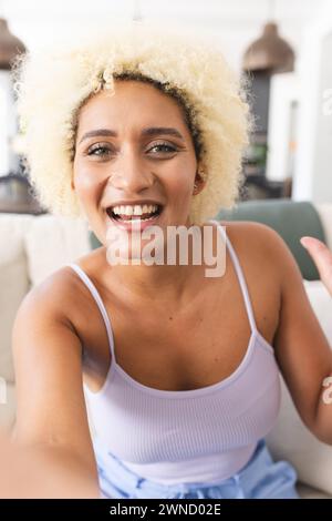 Jeune femme biraciale aux cheveux blonds bouclés sourit à la caméra en appel vidéo Banque D'Images
