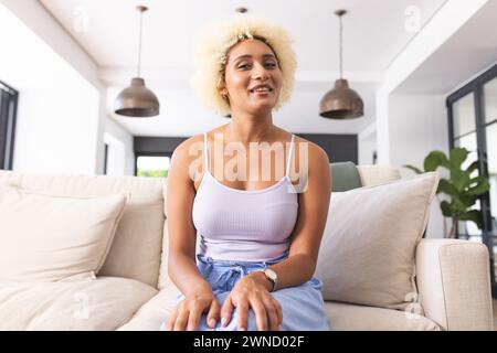 Jeune femme biraciale avec des cheveux bouclés blonds est assise confortablement sur un canapé beige en appel vidéo Banque D'Images