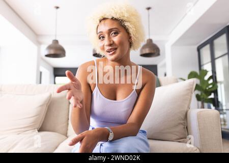 Jeune femme biraciale aux cheveux blonds bouclés étend sa main dans un geste de bienvenue lors d'un appel vidéo Banque D'Images