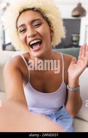 Jeune femme biraciale avec les cheveux blonds bouclés rit joyeusement sur appel vidéo Banque D'Images