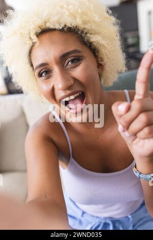 Jeune femme biraciale avec les cheveux blonds bouclés souriante et pointant vers le haut lors d'un appel vidéo Banque D'Images