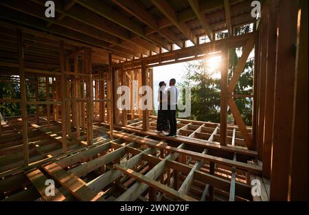 Homme et femme inspectant leur future maison de cadre en bois nichée dans les montagnes près de la forêt. Jeune couple sur le chantier en début de matinée. Concept de construction écologique contemporaine. Banque D'Images