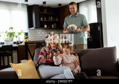 Une famille de trois est confortablement nichée sur un canapé, leurs visages reflétant l'excitation et l'attention comme ils partagent un bol de pop-corn pendant un susp Banque D'Images