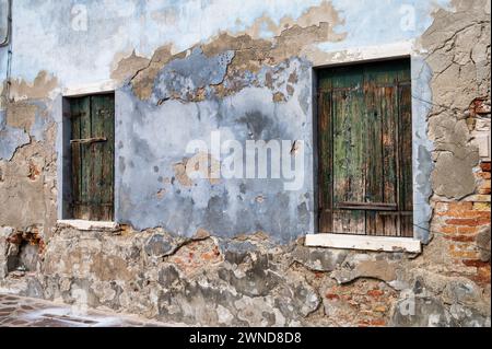 Briques exposées sur un vieux mur de plâtre à Burano Italie avec deux fenêtres à volets en bois. Banque D'Images