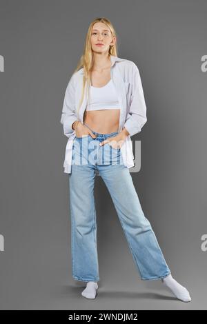 Une femme grande et élancée dans un débardeur, une chemise déboutonnée, un Jean et des chaussettes posant sur un fond gris Banque D'Images