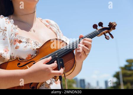 Gros plan d'une jeune violoniste caucasienne en robe florale blanche, debout dans la rue avec son violon et son arc, touchant des cordes avec ses doigts. copier Banque D'Images