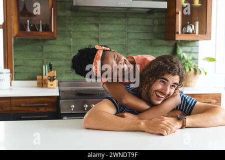 Couple diversifié jeune femme afro-américaine embrasse un jeune homme caucasien par derrière dans une cuisine Banque D'Images
