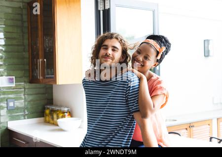 Couple diversifié jeune femme afro-américaine embrasse un jeune homme caucasien dans une cuisine confortable Banque D'Images