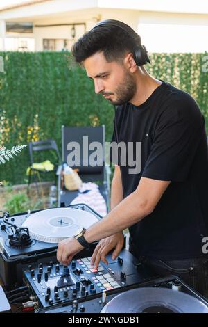 Jeune homme aux cheveux foncés apprenant à utiliser le mixeur dans le jardin de sa maison pour devenir DJ. Banque D'Images