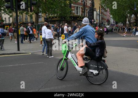 Un homme et une femme passager pilote sur un vélo de location électrique Lime à travers Cambridge Circus, Charing Cross Road, Londres, Royaume-Uni. 1er juillet 2023 Banque D'Images