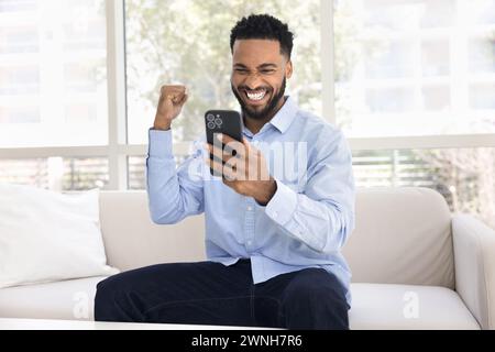 Jeune homme africain couronné de succès tenant un smartphone, faisant la main gagnante Banque D'Images