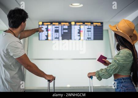de derrière couple de mariage multiculturel à la recherche de leur vol sur l'écran d'information de l'aéroport tout en tenant leurs bagages et passeports Banque D'Images