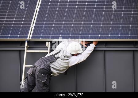 Constructeur connectant les câbles tout en installant des panneaux solaires photovoltaïques sur le toit de la maison. Vue arrière du travailleur dans le montage du casque blanc du module solaire PV sur le toit. Concept d'énergie alternative. Banque D'Images