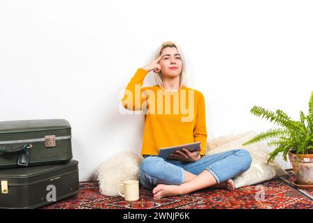 Femme réfléchie dans un pull jaune tient une tablette, touchant sa tempe dans une pose de pensée classique, suggérant la contemplation ou la prise de décision, dans un Banque D'Images