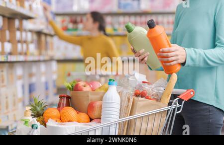 Femme achetant des produits à l'épicerie, elle compare deux bouteilles de détergents Banque D'Images