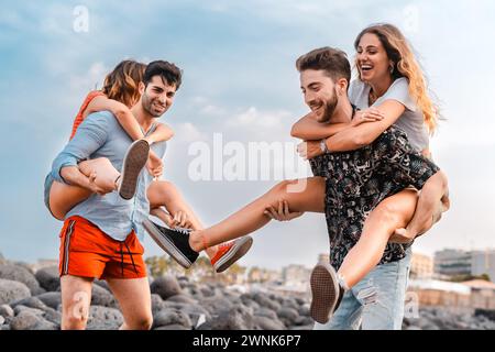 Couple ludique en vacances - Une scène vibrante de deux jeunes couples s'engageant dans des promenades ludiques de piggyback le long de la plage rocheuse, avec des rires remplissant le Banque D'Images