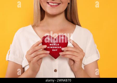 Femme souriante tenant un coeur en bois avec des mots Journée mondiale de la gentillesse sur fond jaune, gros plan Banque D'Images