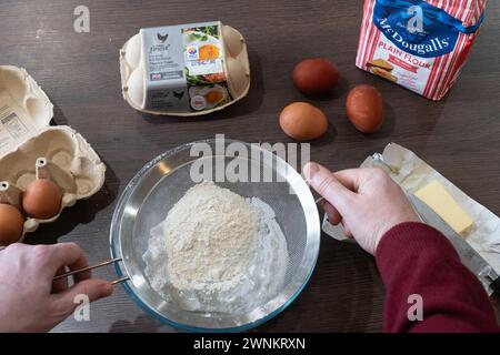 Les mains d'un homme tenant un tamis et tamisant la farine, avec des ingrédients pour cuire un gâteau sur un plan de travail de cuisine - beurre, œufs et farine. ROYAUME-UNI Banque D'Images
