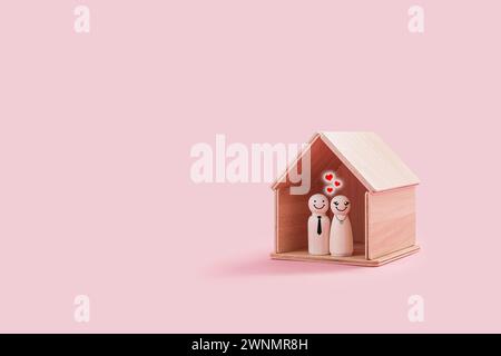 Joyeuses figures masculines et féminines en bois à l'intérieur d'une minuscule maison en bois isolée sur un fond rose. Le couple souriant et se tient proche les uns des autres Banque D'Images