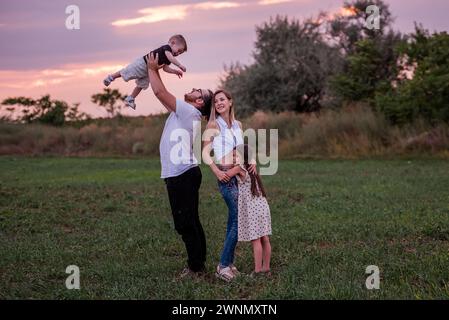 Diversité Happy Family joue sur le terrain au coucher du soleil. Jeune père soulevant le tout-petit haut dans les airs comme la fille aînée et la mère regardent avec charcuterie Banque D'Images