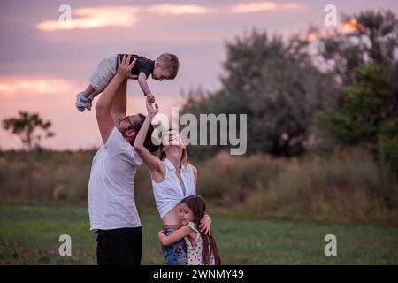 Diversité Happy Family joue sur le terrain au coucher du soleil. Jeune père soulevant le tout-petit haut dans les airs comme la fille aînée et la mère regardent avec charcuterie Banque D'Images