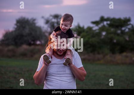 Diversité père avec la barbe portant son fils sur les épaules partage moment tendre dans le champ éclairé crépusculaire, incarnant l'essence de l'amour paternel. Homme et garçon Banque D'Images