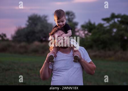 Diversité père avec la barbe portant son fils sur les épaules partage moment tendre dans le champ éclairé crépusculaire, incarnant l'essence de l'amour paternel. Homme et garçon Banque D'Images