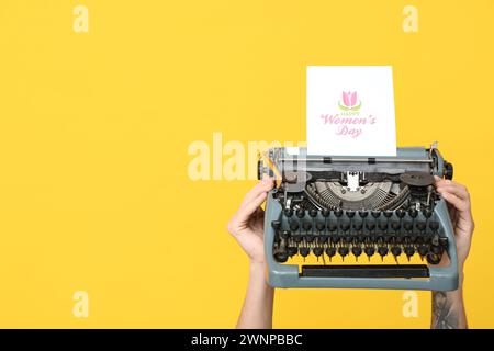 Mains tenant la machine à écrire vintage et la carte postale festive avec le texte HAPPY WOMEN'S DAY sur fond jaune Banque D'Images