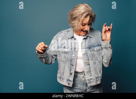 Femme heureuse, mature en denim décontracté se tient dans un studio, souriant et s'amusant tout en dansant. Femme âgée à la mode se déplaçant avec grâce et incarnant Banque D'Images