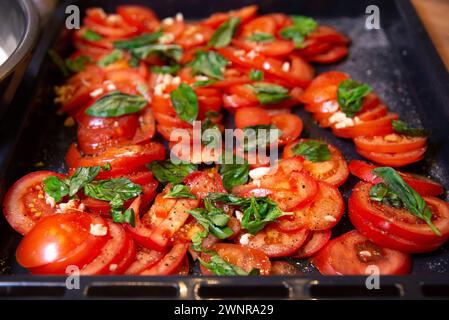 Tranches de tomates mûres assaisonnées d'ail et de poivre, garnies de feuilles de basilic frais sur une plaque de cuisson, prêtes à rôtir. Banque D'Images