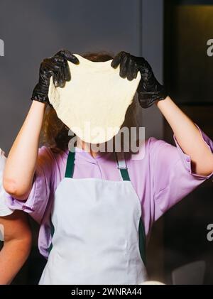 Un chef dans une chemise violette et un tablier blanc étirant la pâte à pizza, portant des gants noirs pour l'hygiène pendant la cuisson. Banque D'Images