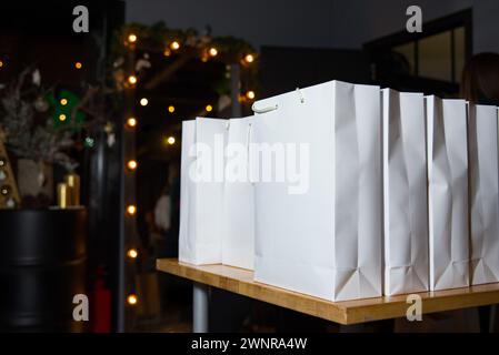 Sacs en papier blanc avec poignées soigneusement alignées sur une table en bois, avec des lumières festives et un décor en arrière-plan. Banque D'Images