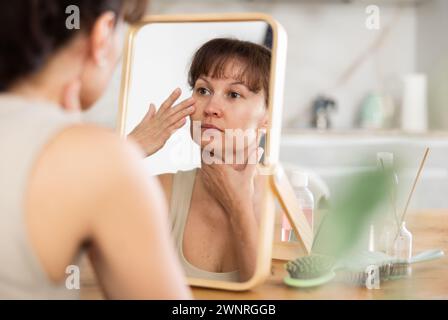 Femme inspectant la peau du visage dans le miroir pendant la routine quotidienne Banque D'Images