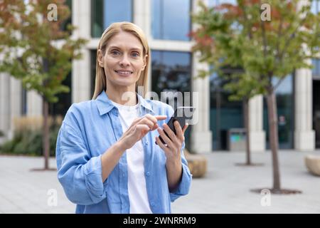 Une jeune femme adulte se tient dehors, concentrée sur son smartphone, avec des bâtiments modernes en arrière-plan. L'image capture une scène commune de connectivité et de style de vie urbain. Banque D'Images