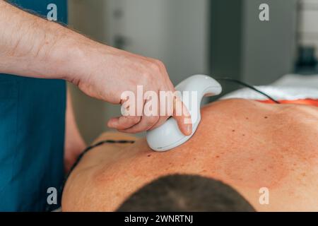Gros plan d'un physiothérapeute appliquant un masseur à percussion sur le dos d'un patient masculin pendant la séance de thérapie Banque D'Images