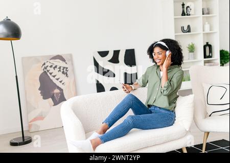 Capturée dans une pièce bien décorée, une femme afro-américaine avec des écouteurs profite d’un moment de loisir avec son smartphone, dégageant une ambiance détendue et tendance Banque D'Images